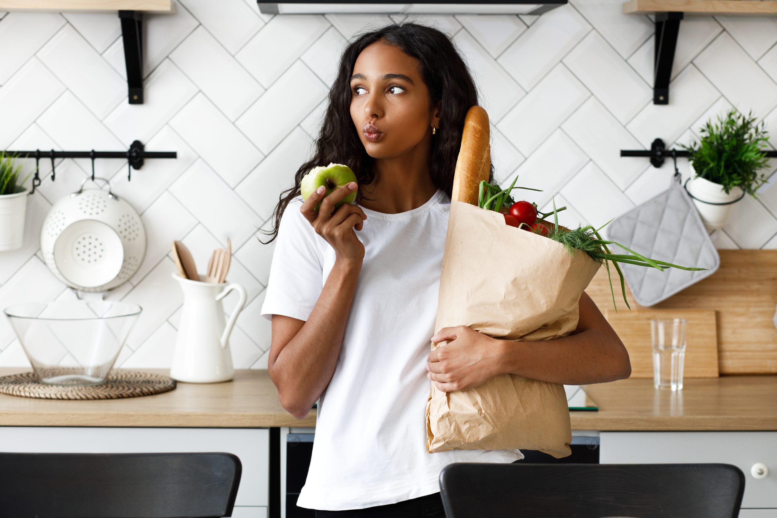 Thoughtful mulatto girl is holding package full with fresh vegetables in one hand and bitten apple in other, on the modern white kitchen