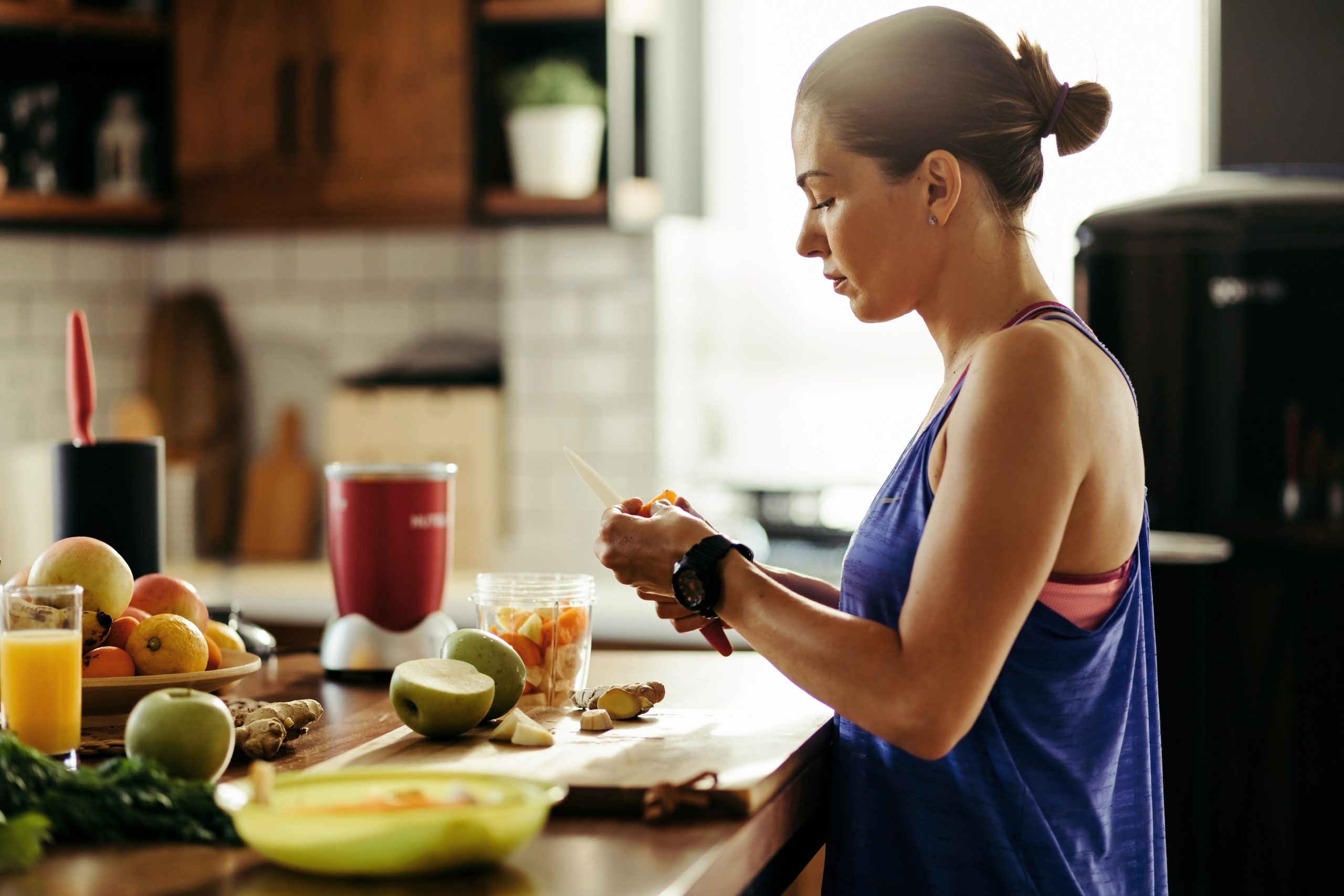 Young sportswoman making herself a healthy smoothie and slicing fresh fruit in the kitchen.